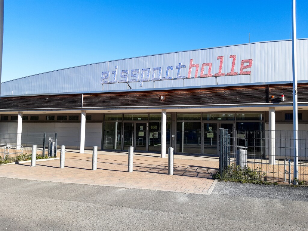 Das Foto zeigt die Außenansicht einer Halle mit der Aufschrift Eissporthalle und einen gepflasterten Vorplatz.
