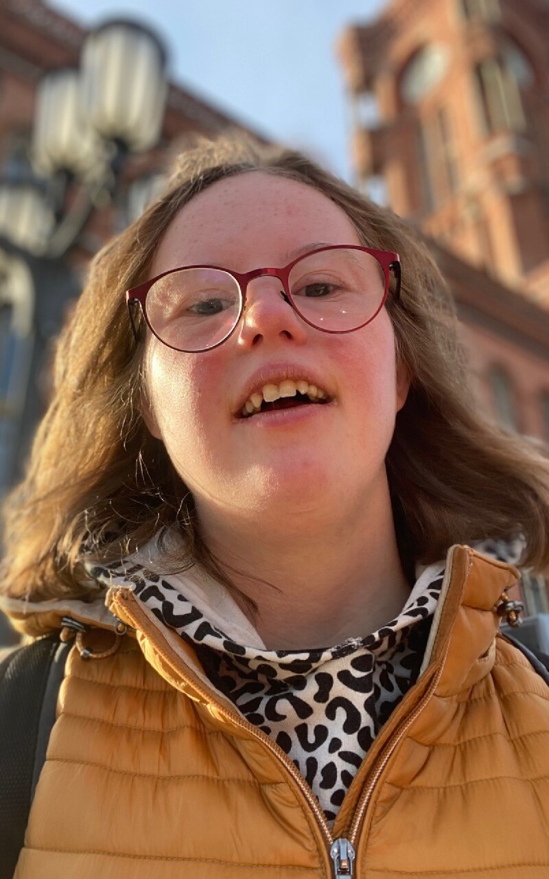 Auf dem Foto lächelt Natalie Dedreux, deutsche Aktivistin, Journalistin und Bloggerin, die sich als Betroffene für die Inklusion von Menschen mit Down-Syndrom engagiert, in die Kamera. Sie trägt eine senfgelbe Jacke und eine rote Brille.