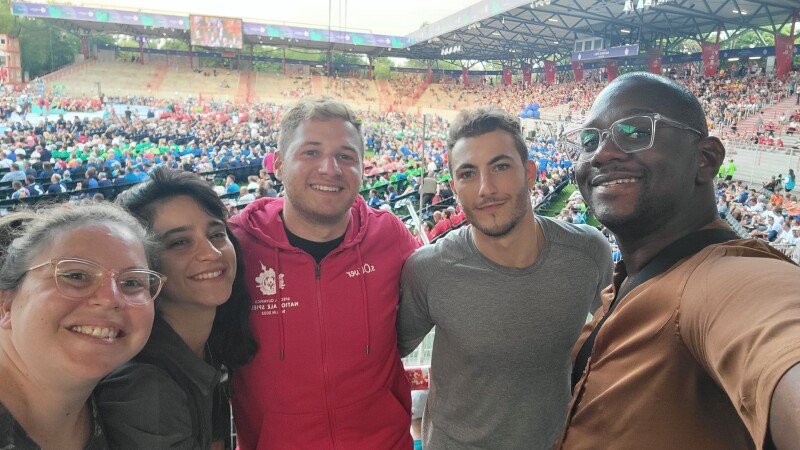 Das Foto ist ein Selfie, aufgenommen während der Eröffnungsfeier und von einem Corporate Volunteer. Er posiert gemeinsam mit 4 weiteren Volunteers und alle lächeln fröhlich in die Kamera.