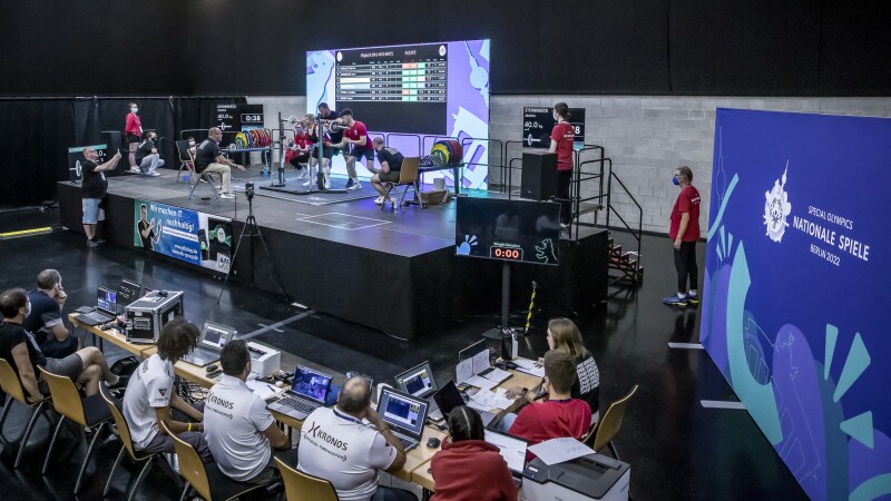 In the photo you can see the competition place of the powerlifting competition. 8 people are sitting at tables. Among them are Kronos officials and the competition management. On the stage you can see judges and athletes getting ready. Volunteers are standing on the right.