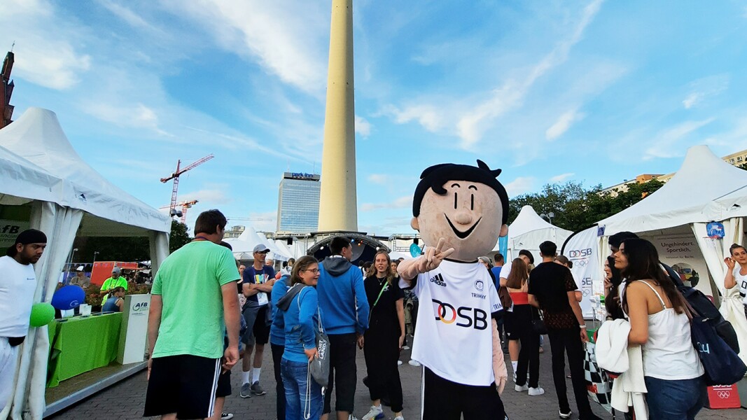 The photo shows the Berlin TV Tower in the background and some people and stands of the Special Olympics Festival in the foreground. In addition, the mascot of the DOSB poses in front of it.