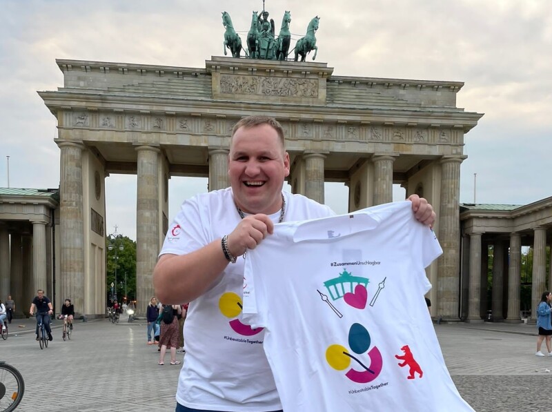 The photo shows athlete spokesperson Dennis Mellentin standing in front of the Brandenburg Gate, both wearing the newly designed T-shirt and laughing as he holds it up to the camera.