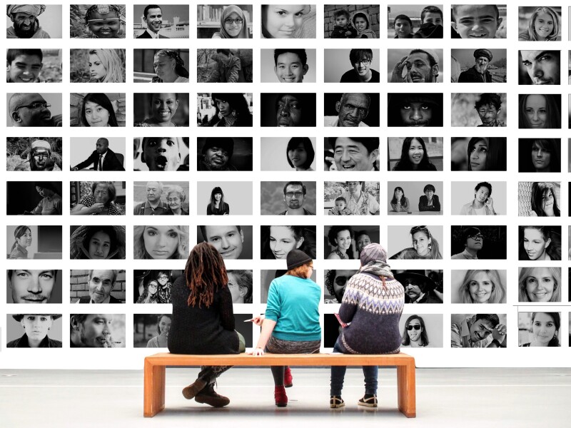 The photo shows a scene in a museum or exhibition. Three people sit on a bench and look at a wall full of pictures.