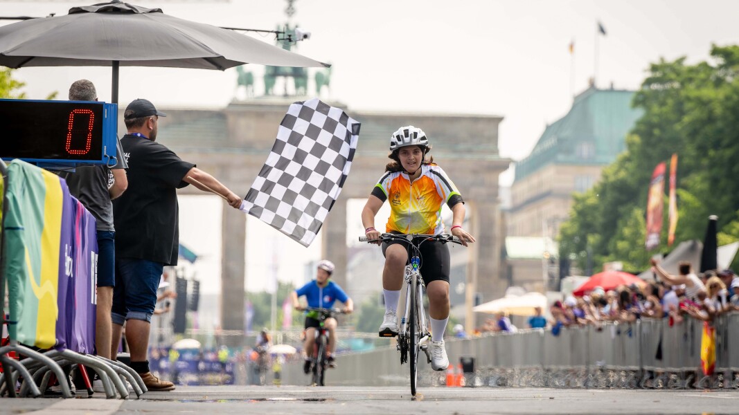 Das Foto zeigt eine griechische Radsportlerin beim Zieleinlauf des Straßenrennens mit dem Brandenburger Tor im Hintergrund. Sie tritt noch einmal kräftig in die Pedale und sieht dabei sehr glücklich aus, gleich das Ziel erreicht zu haben.