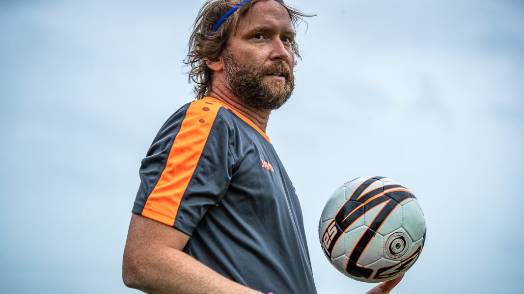 In the photo there is a soccer player who is holding the ball in his hand. He is wearing a gray t-shirt and looks concentrated.