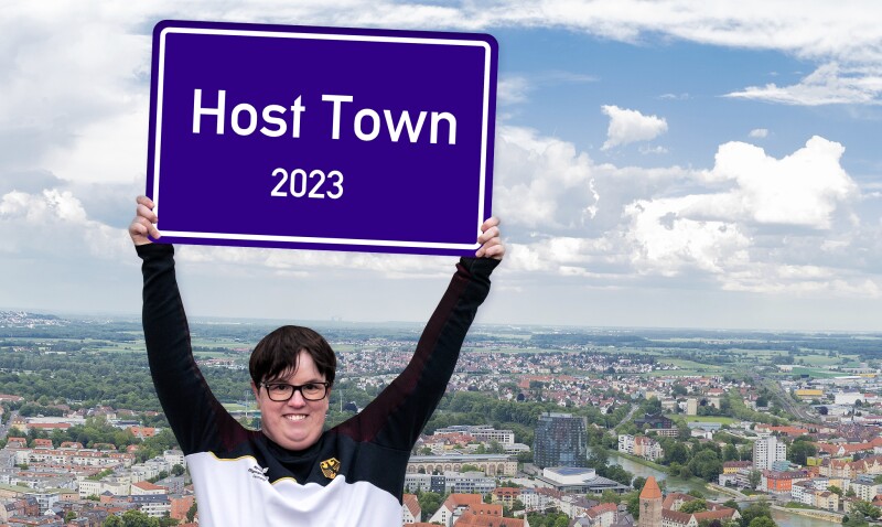 Special Olympics athlete Juliana Rößler holds a sign with the words Host Town in the air. Behind her, you can see a town.