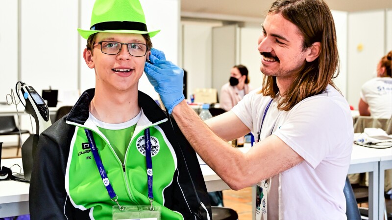 Auf dem Foto sieht man eine Healthy Hearing Untersuchung im Rahmen des Healthy Athletes Programms. Ein Arzt untersucht gerade das Ohr eines Athleten, der entspannt in die Kamera lächelt.