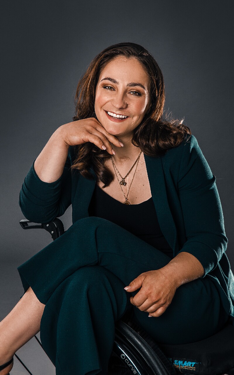 The photo shows the Olympic Champion in track cycling Kristina Vogel. She is sitting in her wheelchair with her legs crossed and smiling, wearing a black blazer.