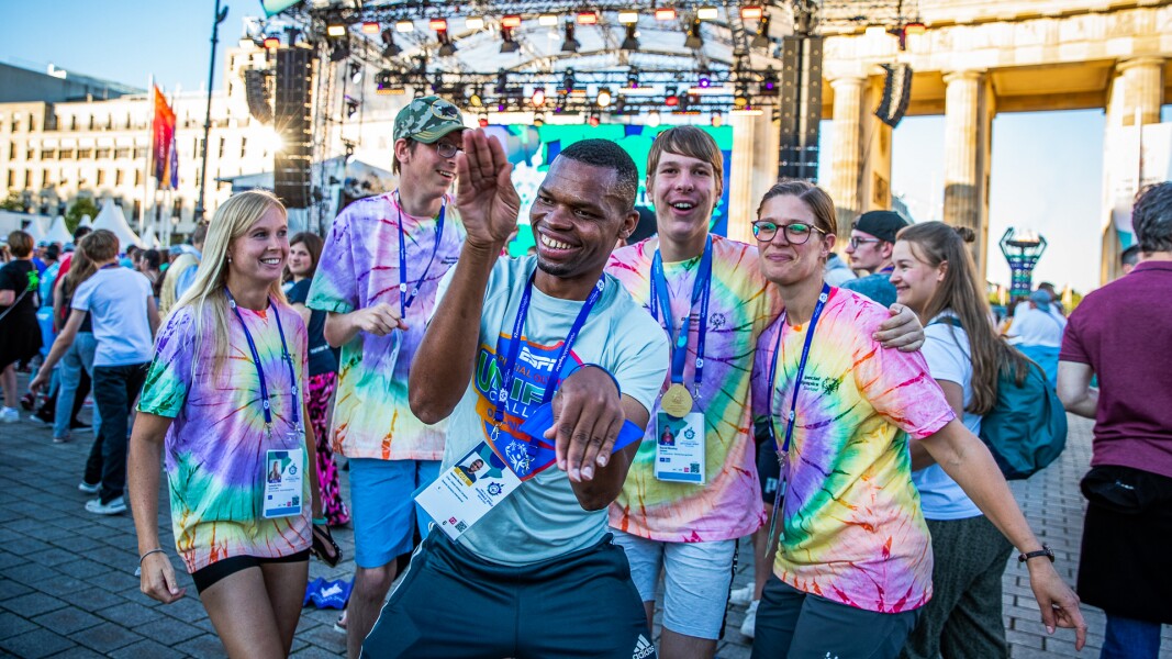Auf dem Foto tanzt eine Athletengruppe von fünf Personen ausgelassen und lächelnd vor der Bühne und dem Brandenburger Tor.