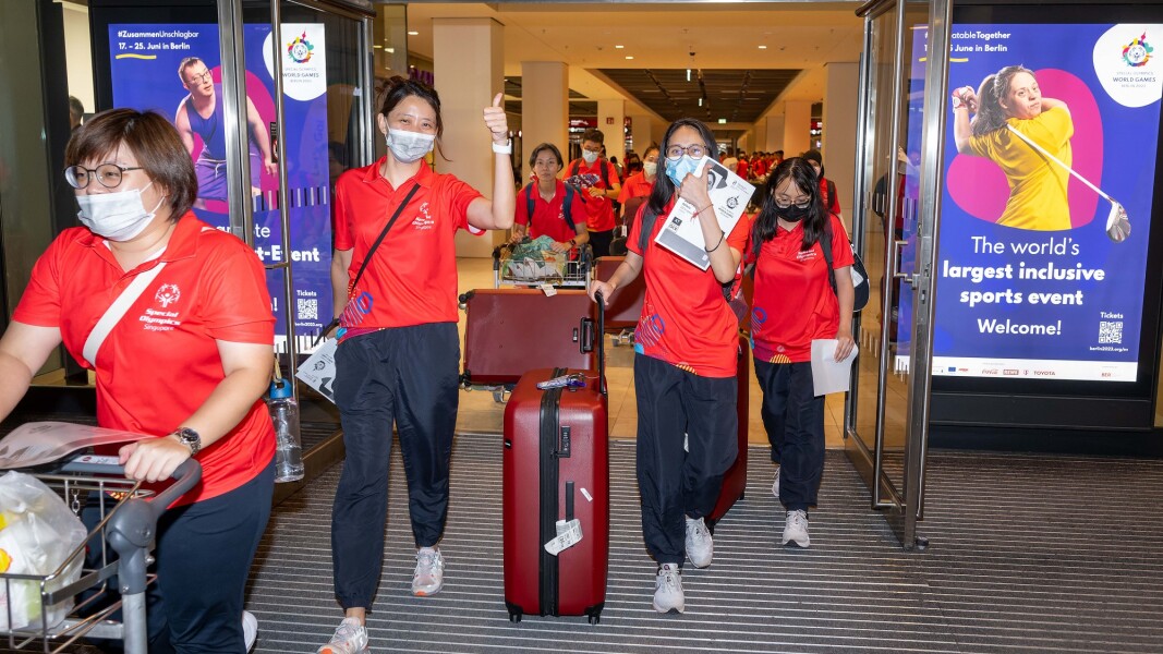 The photo shows the arrival of the athletes from Singapore, who are just coming out of the airport through the doors with their luggage, looking very happy.