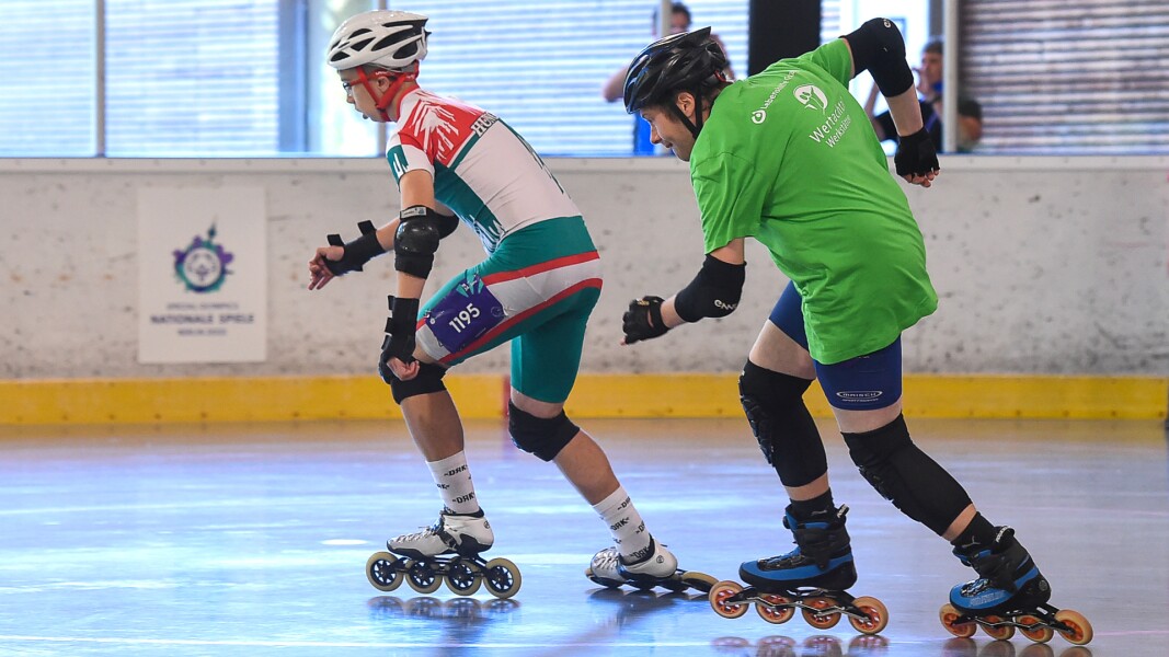Auf dem Foto sieht man zwei Roller Skater in ihrem Wettbewerb. Der hintere versucht den vorderen aufzuholen.