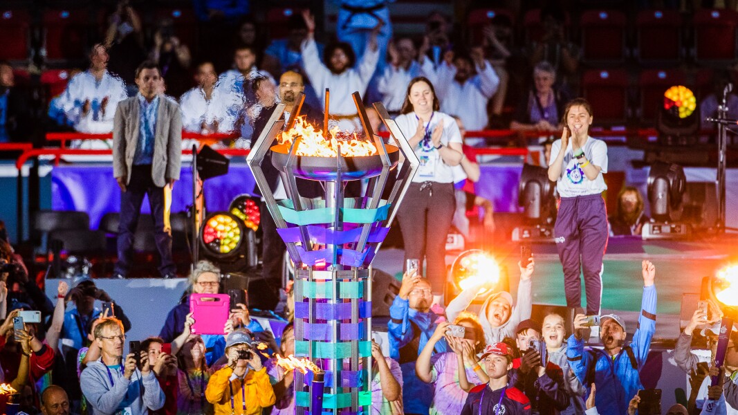 The photo shows a scene of the Opening Ceremony of the National Games in Berlin. In the center is the big torch flame and around it are some people taking photos of it while cheering.
