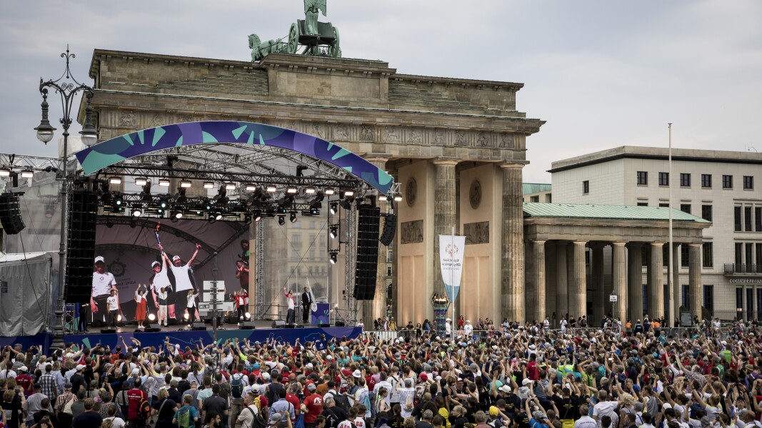 The photo shows the Closing Ceremony of the National Games in Berlin. A large stage is set up directly in front of the Brandenburg Gate and hundreds of people celebrate together.