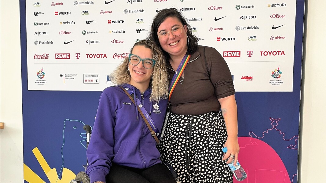 Das Foto zeigt zwei Frauen, die sich vor einer Wand mit den Sponsoren der Special Olympics World Games 2023 Berlin fotografieren lassen. Sie lächeln freundlich in die Kamera.