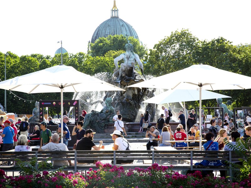 Das Foto zeigt eine Szene des Special Olympics Festivals direkt am berühmten Neptunbrunnen neben dem Alexanderplatz im Herzen Berlins. Viele Menschen schlendern entweder vorbei oder sitzen entspannt auf Bänken.