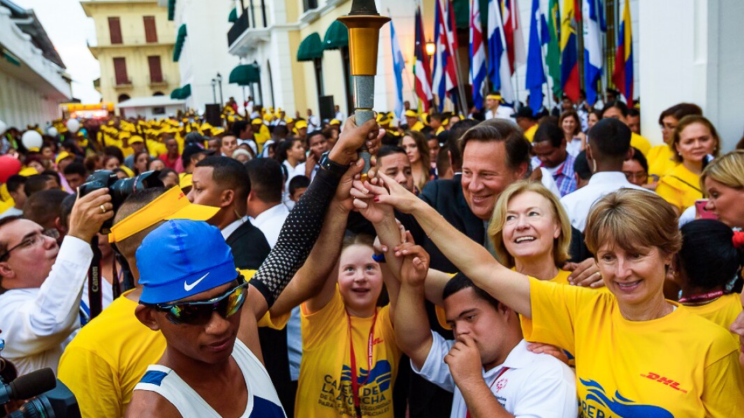 The photo shows an athlete with the Special Olympics torch in his hand. Around him are many people who are also touching the torch and smiling.