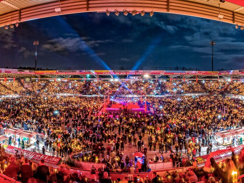The photo shows the Stadion An der Alten Försterei filled with many people to a major event. The colors are bright in front of the evening sky.