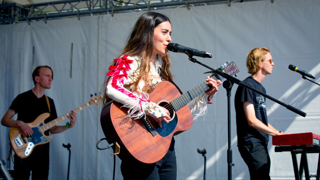 In the picture you can see three musicians+*. In the foreground there is a singer with her guitar. In the background you can see two men. One of them is playing the keyboard and the other one is also playing the guitar.