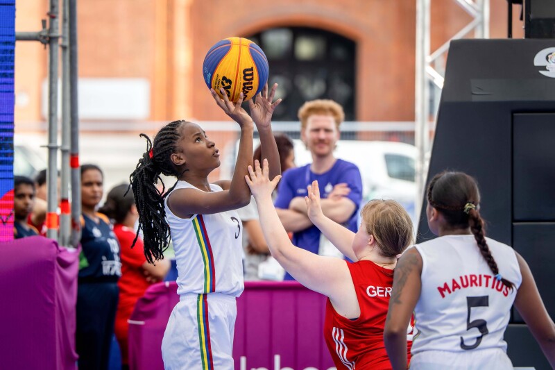 The Mauritian female athletes wearing the white jerseys is trying to score while the German female athlete wearing the red jersey is trying to block the shot