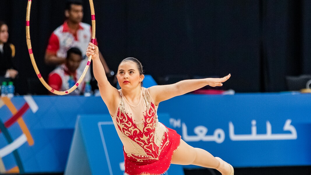 Das Foto zeigt eine Szene einer Athletin bei der Rhythmischen Sportgymnastik. Sie hebt einen Reifen und ein Bein.