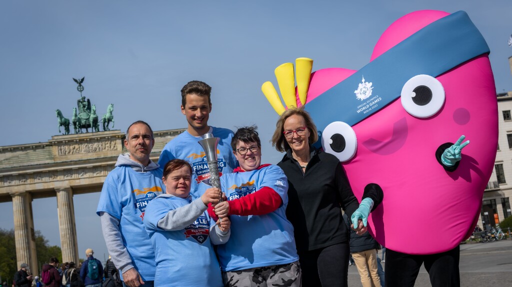 Auf dem Foto sind Athlet*innen und das Maskottchen Unity und Christiane Krajewski zu sehen. Sie präsentieren freudig die Flacke.