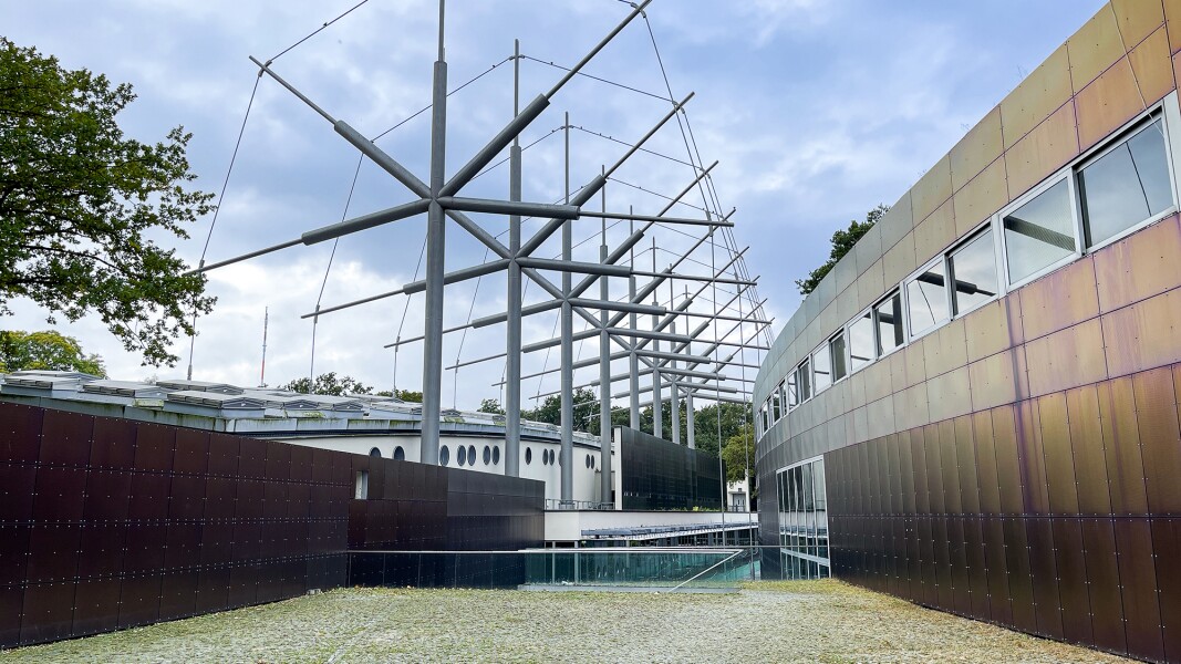 Das Foto zeigt ein weißes Gebäude im Hintergrund und ein Gebäude mit einer Metallfassade im Vordergrund. In der Mitte zwischen beiden Gebäuden ist eine Stahlkonstruktion zu sehen.