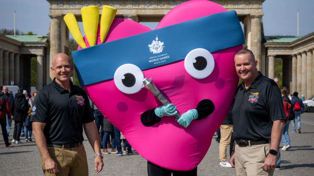 Auf dem Foto sind zwei Polizisten und das Maskottchen Unity zu sehen. Unity hält die Fackel.