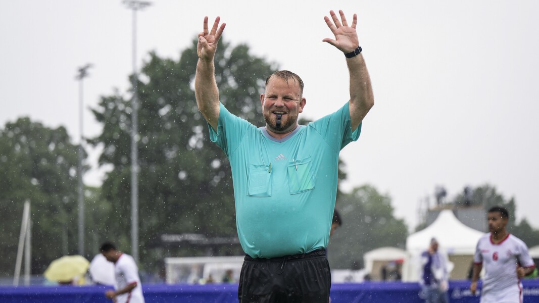 The photo shows a referee playing soccer. He is giving hand signals and smiling with his whistle in his mouth. It is raining, but he is still in a good mood.