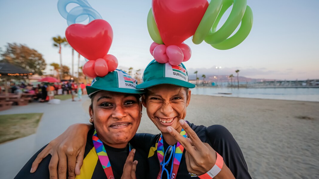 In the photo there are two women on the beach laughing and forming the peace sign with their fingers.