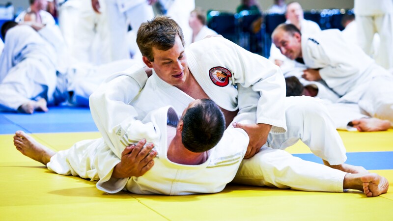 Auf dem Foto treten zwei männliche Judoka miteinander an. Der eine liegt mit dem Rücken auf dem Boden und der andere drückt ihn Richtung Boden. Im Hintergrund sind weitere Judoka Paare zu sehen, die miteinander antreten und trainieren.