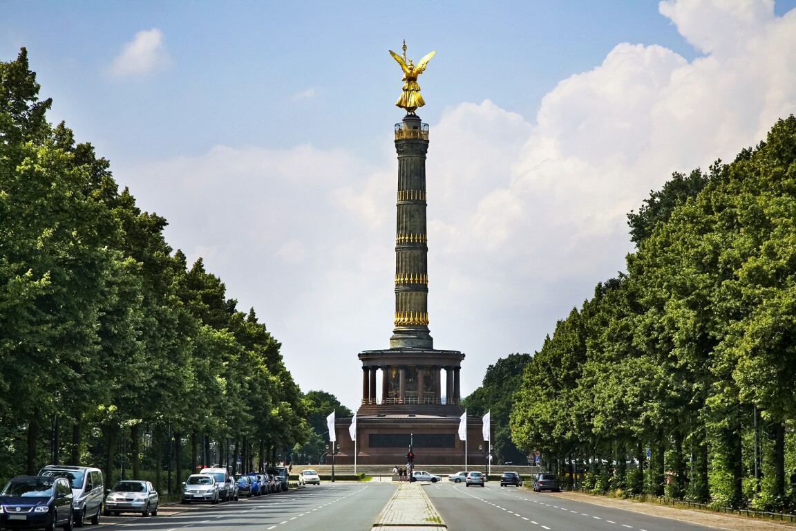 The photo shows a street with parked cars and in the center the Victory Column in Berlin.