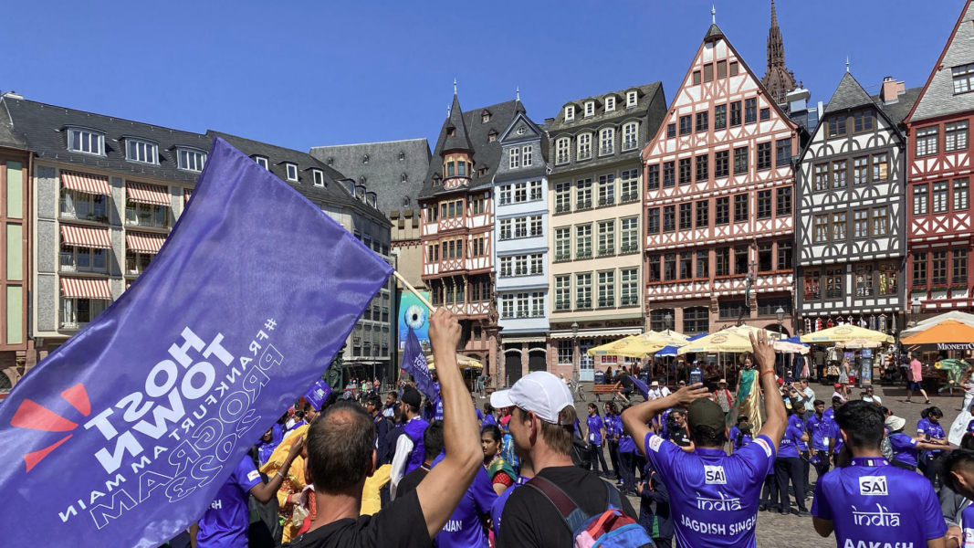 The photo shows the Indian delegation waving the Host Town flag at the Rathausplatz in Frankfurt. Half-timbered houses can be seen in the background.