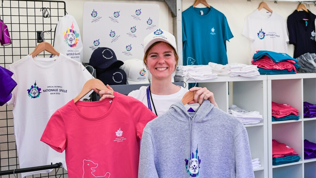 Auf dem Foto ist eine Mitarbeiterin im Merchandise-Stand zu sehen, die verschiedene Artikel lächelnd hochhält. Der Stand ist Teil des Special Olympics Festivals.