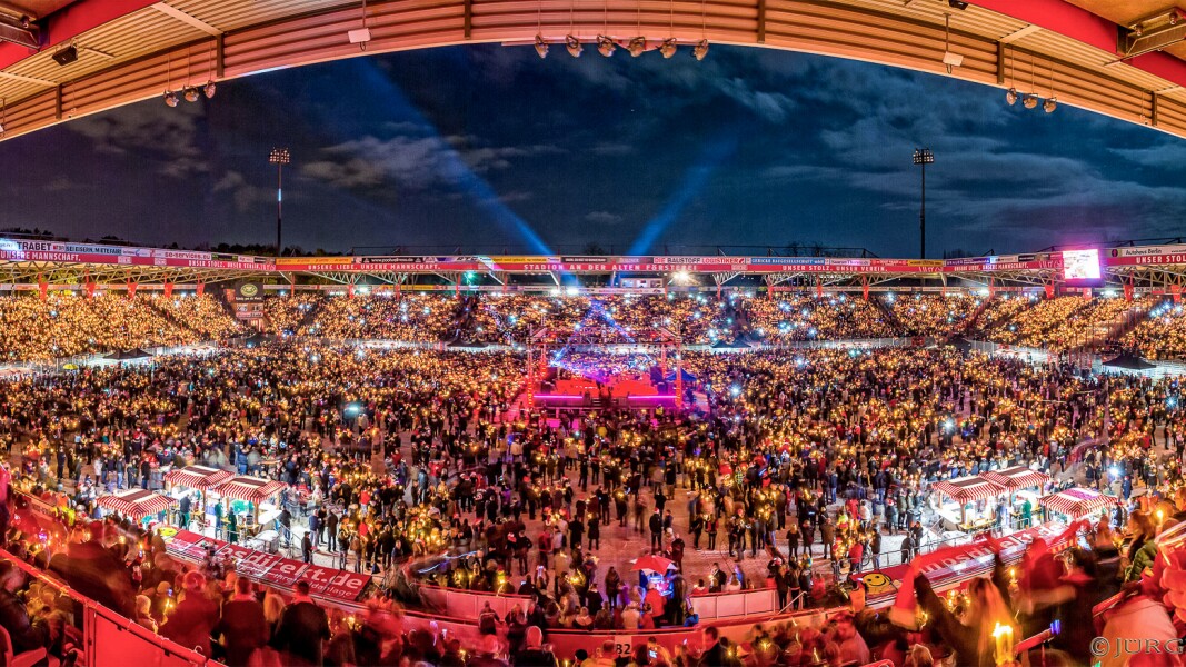 The photo shows the Stadion An der Alten Försterei filled with many people to a major event. The colors are bright in front of the evening sky.