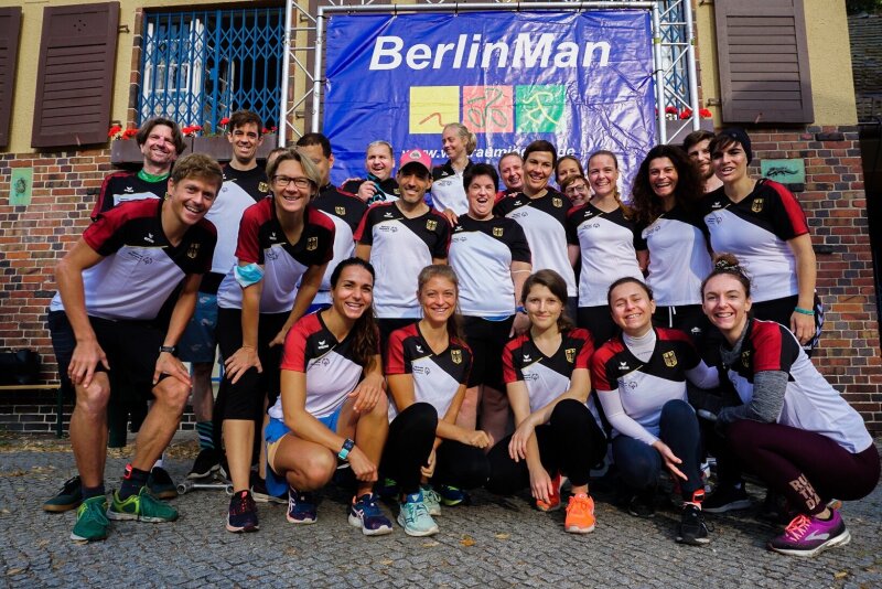 Alle+Teilnehmer_innen+des+Triathlons+posieren+gemeinsam+und+mit+Freude+vor+dem+blauen+BerlinMan-Banner