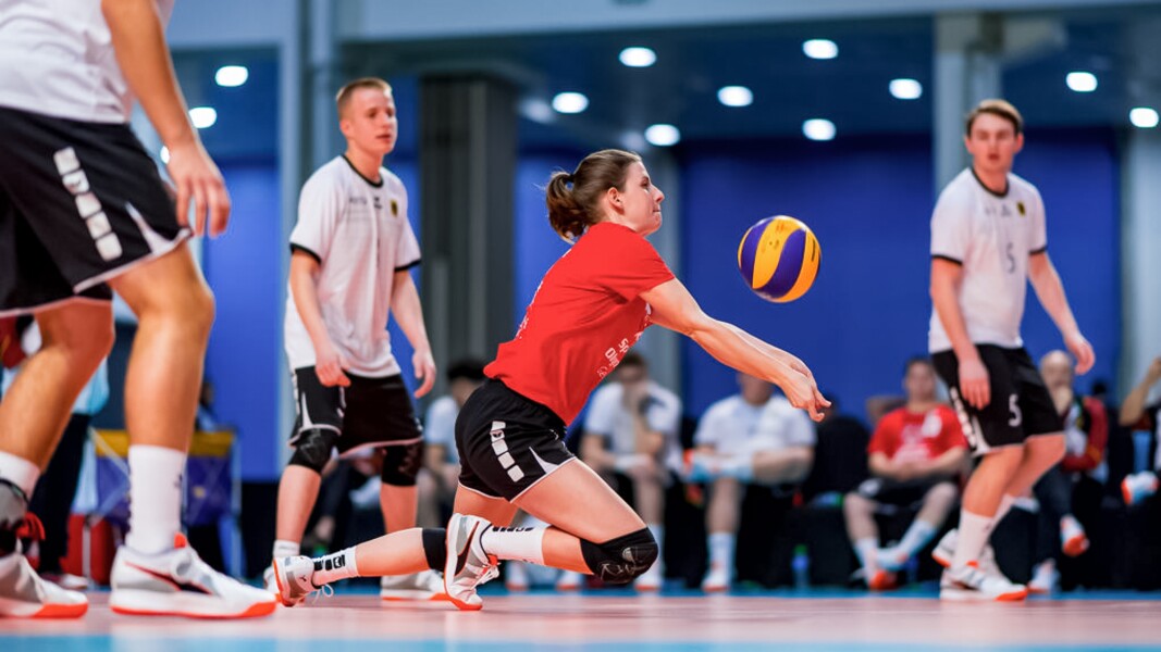 Das Foto zeigt eine Mannschaft beim Volleyball. Eine Spielerin nimmt gerade den Aufschlag des Gegners an. Sie ist mit ihren Knien fast auf dem Boden.