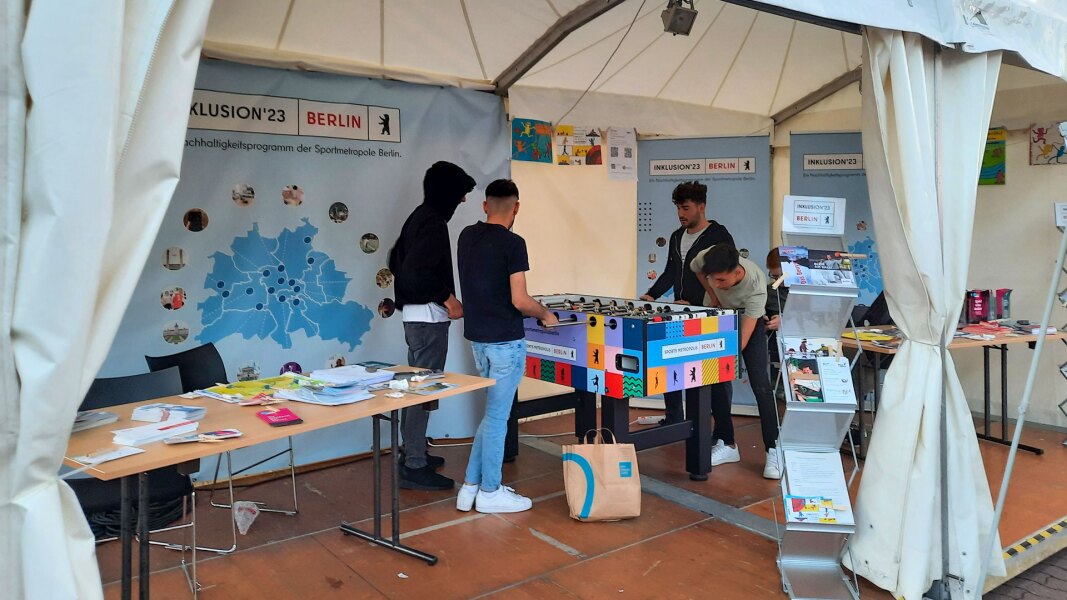 Das Foto zeigt den Aktionsstand von Inklusion '23, einem Inklusionsprojekt der Stadt Berlin. Hier sind gerade vier Jugendliche an einem Kickertisch.