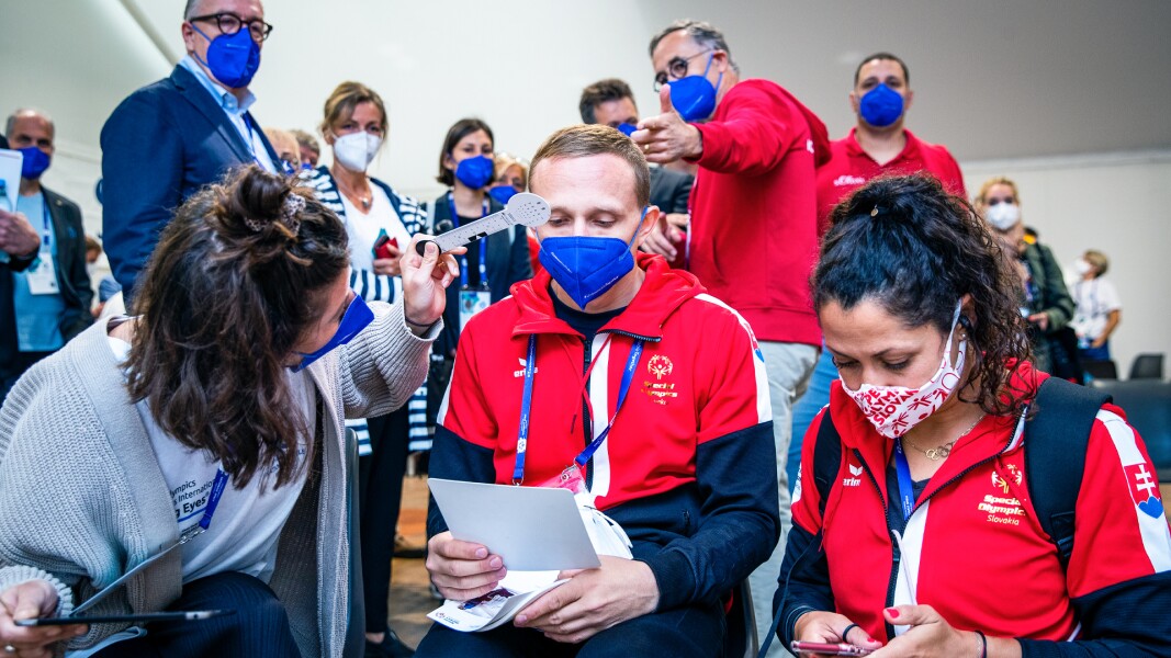 A woman is doing an eye examination with an athlete. Another woman sits next to them and in the background you can see further people watching.