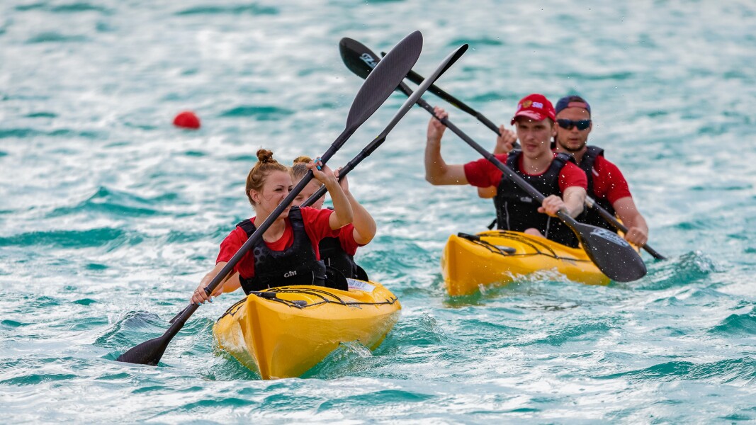 The photo shows two kayaks with two athletes each during competition. On one boat are two women and on the other one two men.