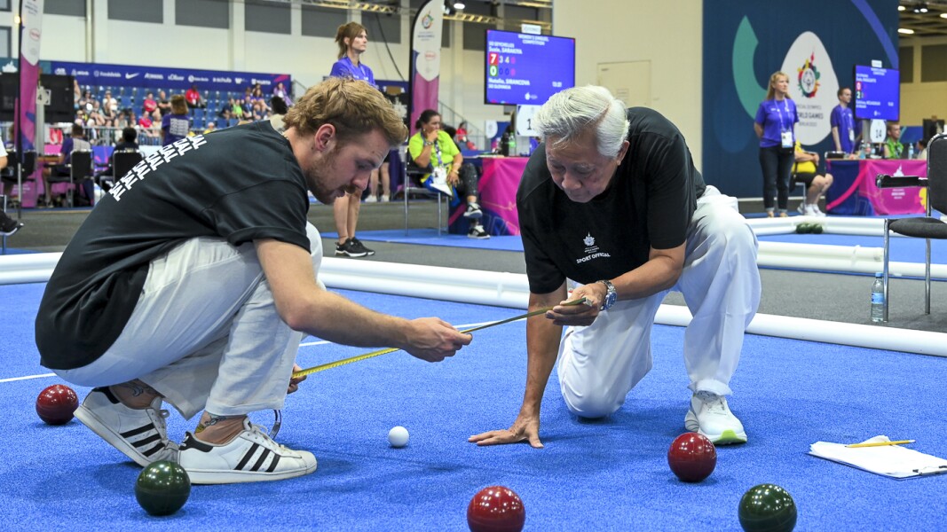 The picture shows two referees playing bocce. They squat on the floor and measure the result with a tape measure.