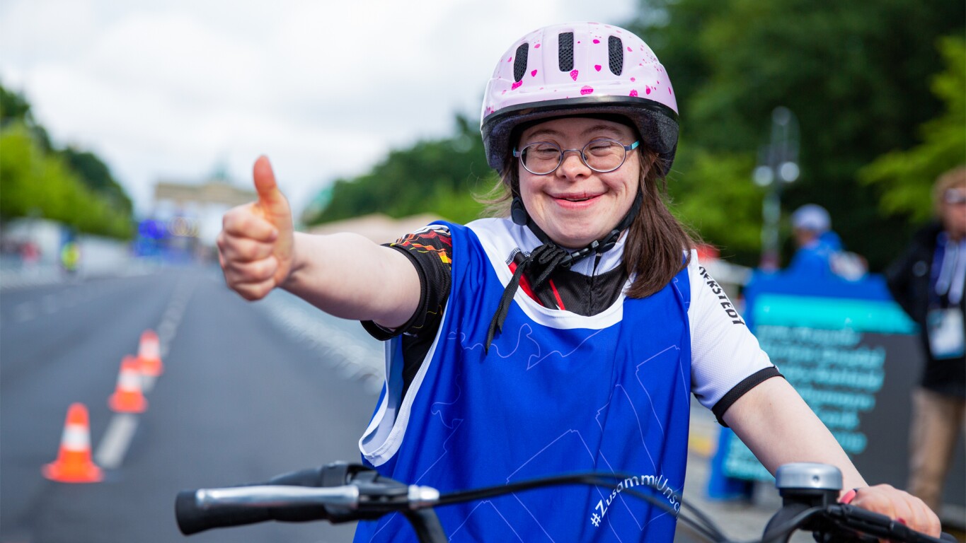 The photo shows a female athlete during her cycling competition at the National Games Berlin 2022. She smiles and shows thumbs up.