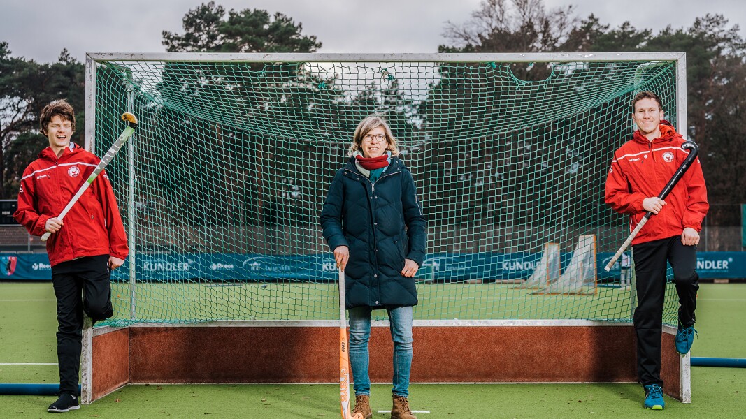 Auf dem Foto sind drei Personen zu sehen, die an einem Hockeytor lehnen und jeweils einen Schläger in der Hand halten.