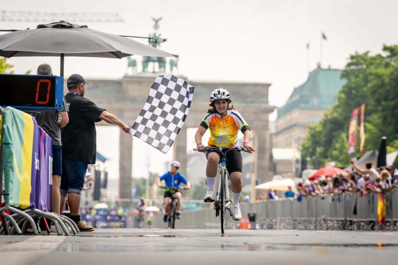 The photo shows a Greek cyclist crossing the finish line of the road race with the Brandenburg Tor in the background. She is  looks very happy to have reached the finish line right away.