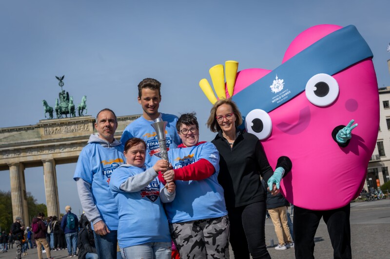 Auf dem Foto sind Athlet*innen und das Maskottchen Unity und Christiane Krajewski zu sehen. Sie präsentieren freudig die Flacke.