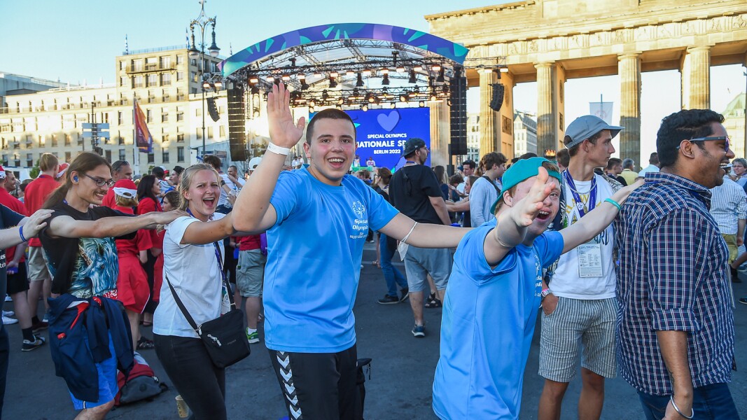 Auf dem Foto sieht man Feiernde, die in einer Polonaise durchs Bild tanzen. Sie lachen und winken. Im Hintergrund ist die Special Olympics Bühne und das Brandenburger Tor zu sehen.