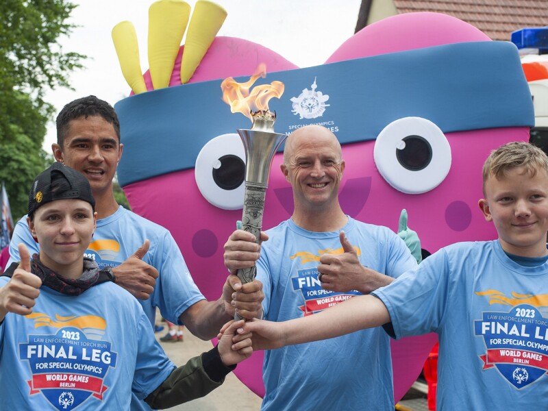 In the photo, the torch is carried by four young kids, who are accompanied by the mascot, a local representative wearing a gray suit, and other people in the background.