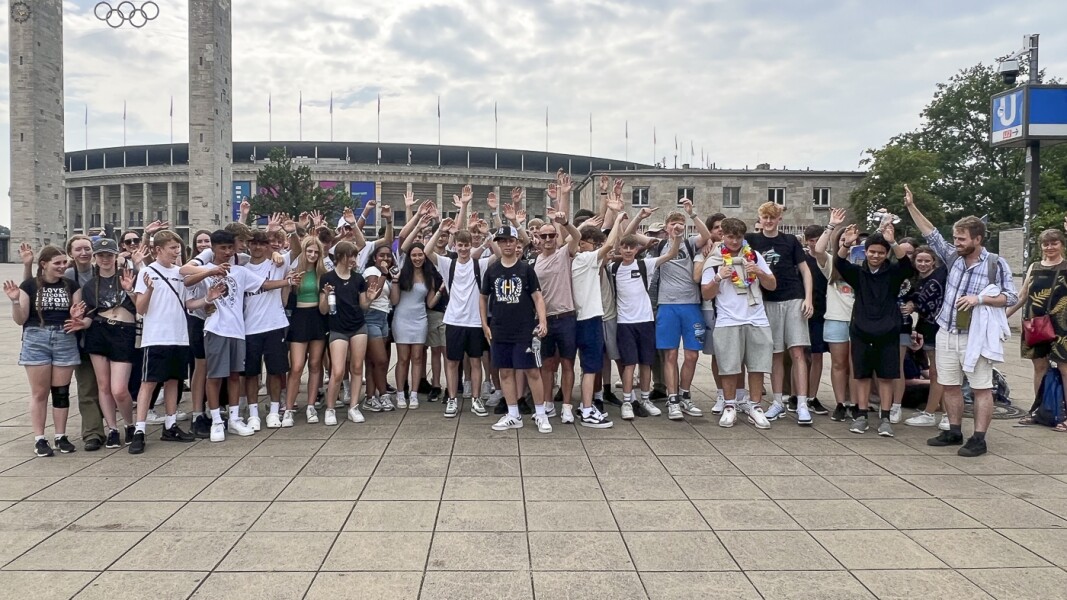 The photo shows a school class in front of the Olympic Stadium. They joyfully stretch their hands up in the air.