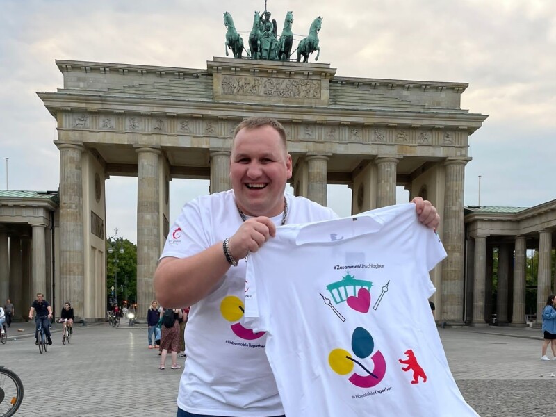 The photo shows athlete spokesperson Dennis Mellentin standing in front of the Brandenburg Gate, both wearing the newly designed T-shirt and laughing as he holds it up to the camera.