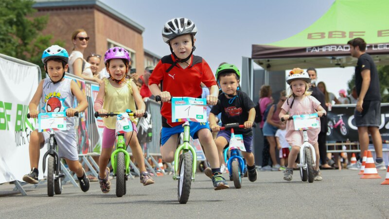 Das Foto zeigt eine Gruppe Kinder, die auf Laufrädern ein Wettrennen fahren. Alle tragen Helme und haben Spaß. Das Bild entstand während des Familiensportfests in Berlin. 