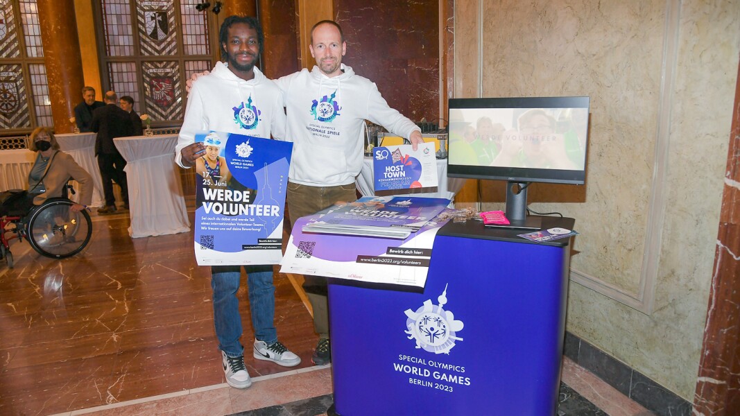 Auf dem Foto sind zwei Mitarbeiter des LOC zu sehen, die Plakate zum Thema "werde Volunteer" hochhalten. Sie sind Teil des Volunteer-Teams und wollen Menschen für diese Arbeit bei Special Olympics gewinnen.
